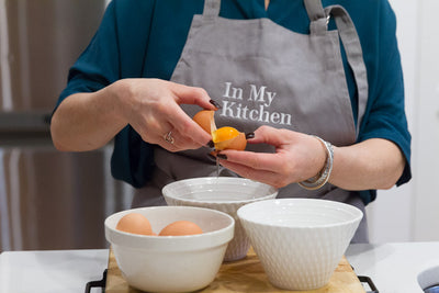 In my kitchen Apron being worn while breaking an egg over a white bowl in a kitchen
